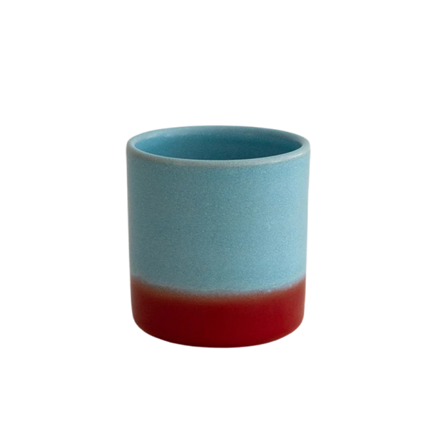 A small cylindrical ceramic cup with a two-tone glaze, featuring a light blue upper section and a deep red lower section, set against a plain white background.